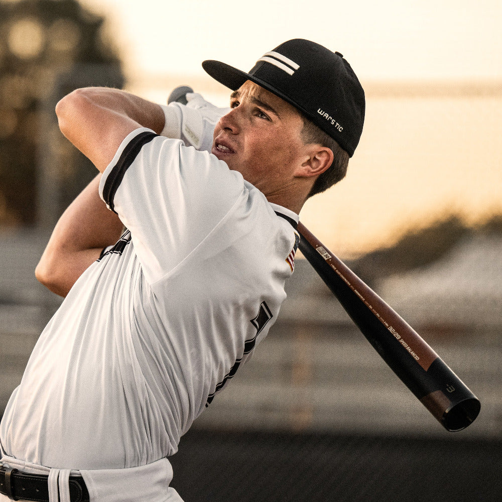 A young player in a white uniform and black cap swings the 2023 Warstic Warhawk3 (-3) BBCOR Baseball Bat (USED), known for its balanced swing and high-performance alloy, near a fence with a blurred outdoor background.