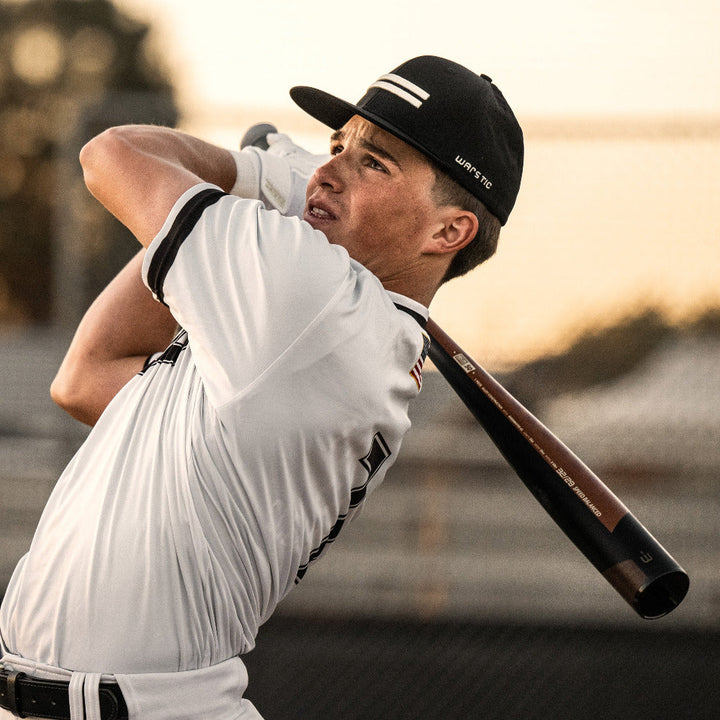 A young player in a white uniform and black cap swings the 2023 Warstic Warhawk3 (-3) BBCOR Baseball Bat (USED), known for its balanced swing and high-performance alloy, near a fence with a blurred outdoor background.
