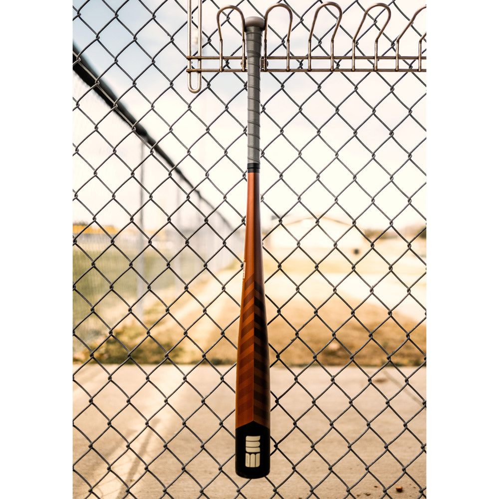 A used 2023 Warstic Warhawk3 (-3) BBCOR Baseball Bat (MBWH323WF3) with a balanced swing weight hangs vertically on metal hooks of a chain-link fence, overlooking an empty outdoor sports field on a sunny day.