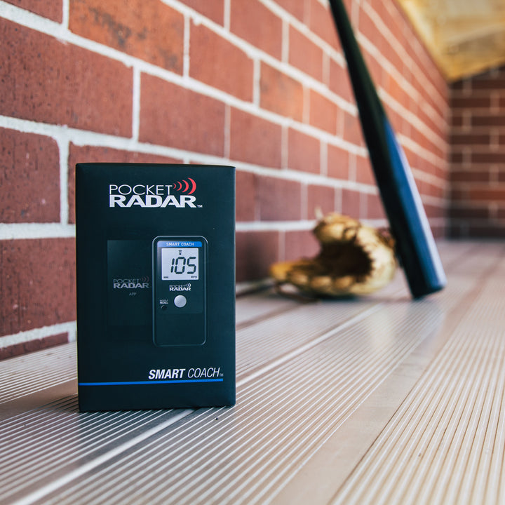 A Pocket Radar Smart Coach Radar: SR1100 by Pocket Radar rests on a metal bench, with a baseball glove and bat blurred in the background—ideal for athletes training with this speed system and the Pocket Radar app.