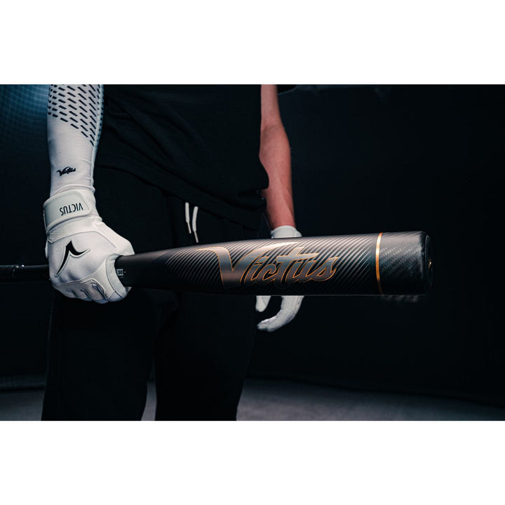 Wearing white batting gloves, a person holds a Victus Vandal 2 (-8) USSSA Baseball Bat (2022, black/gold) with one hand in a dark setting; only their upper body is partially seen.