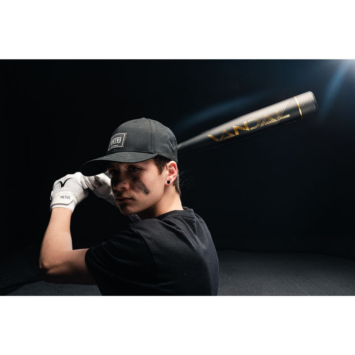 A person with black face paint, dressed in a black cap and shirt with white gloves, poses against a dark background holding a 2022 Victus Vandal 2 (-8) 2 3/4" USSSA Baseball Bat (VSBV2X8) by Victus over their shoulder, ready to swing.