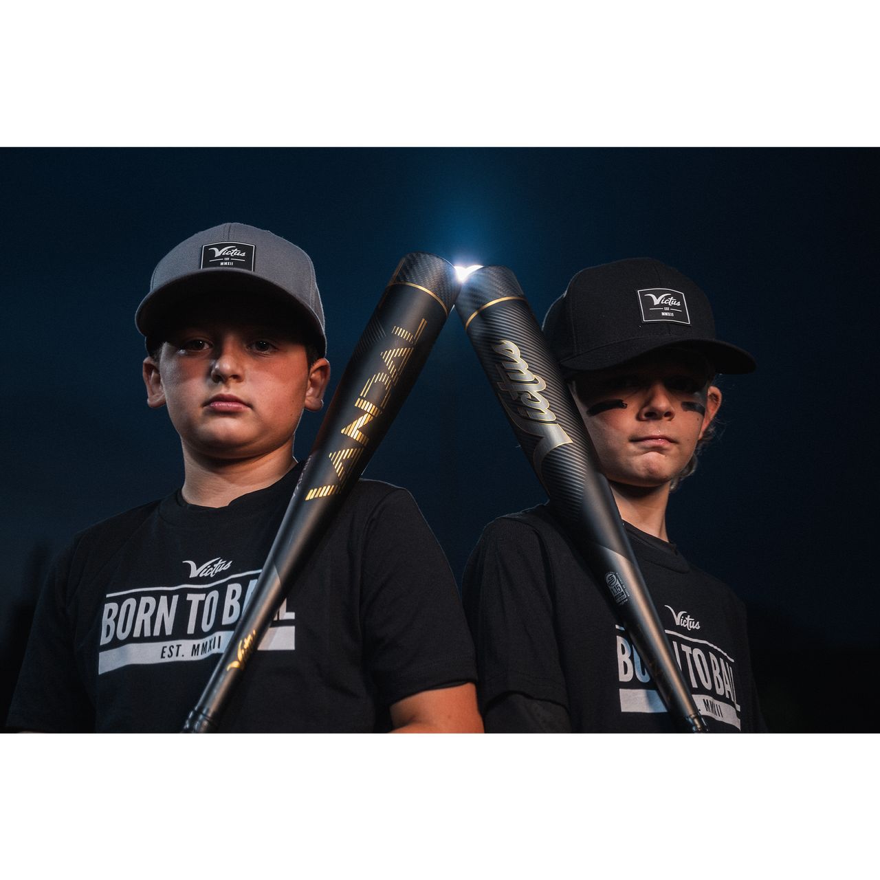 Two boys in black shirts and hats stand side by side at night, holding Victus 2022 Vandal 2 (-8) USSSA Baseball Bats (VSBV2X8), crossed in front of them as the bats shine under a bright light, their serious expressions adding intensity.