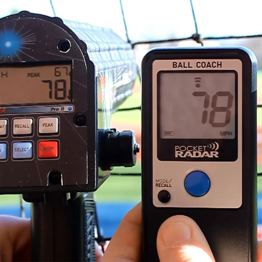 A hand holds the Pocket Radar Ball Coach Radar PR1000-BC by Pocket Radar, displaying 78 MPH beside a larger ball speed device, both positioned in front of a chain-link fence at an outdoor sports field.