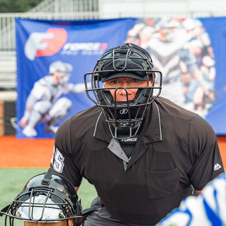 An umpire in a Force3 Pro Gear Traditional Defender Mask: BD31 crouches behind the catcher on the field, with blurred promotional banners and stands in the background.