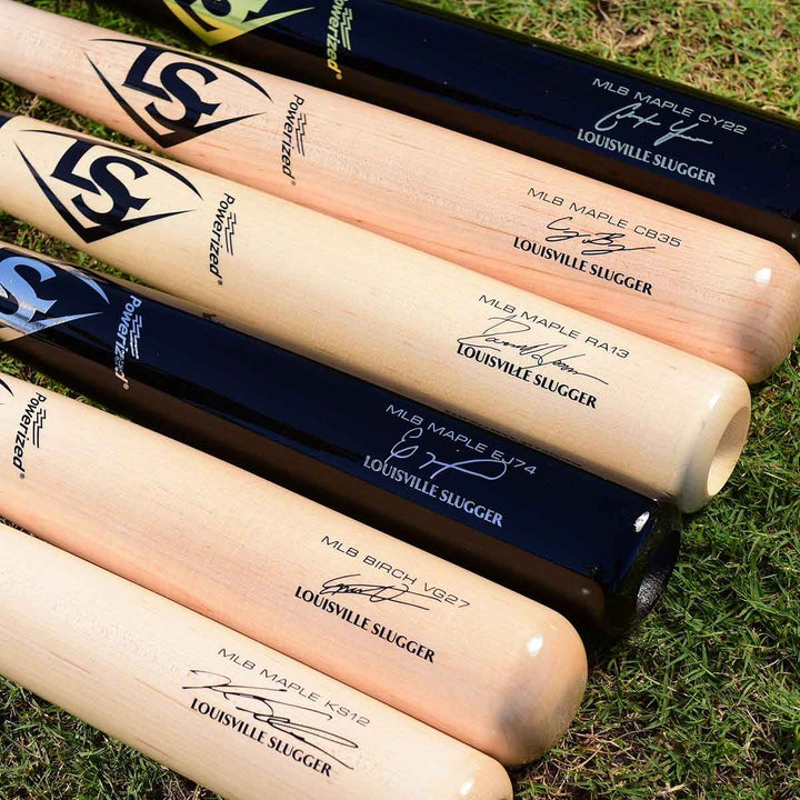 Several Louisville Slugger bats, including the MLB Prime Signature Series CY22 Christian Yelich Game Model (WBL2435010), are arranged side by side on grass, each showing its model number and signature on the barrel.