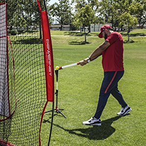 Wearing a red shirt, cap, and dark pants, a person hits a baseball off a tee toward a practice net using PowerNet 3.2" Weighted Hitting and Batting Training Balls (6 Pack): 1004L by PowerNet on a grassy field with trees behind.