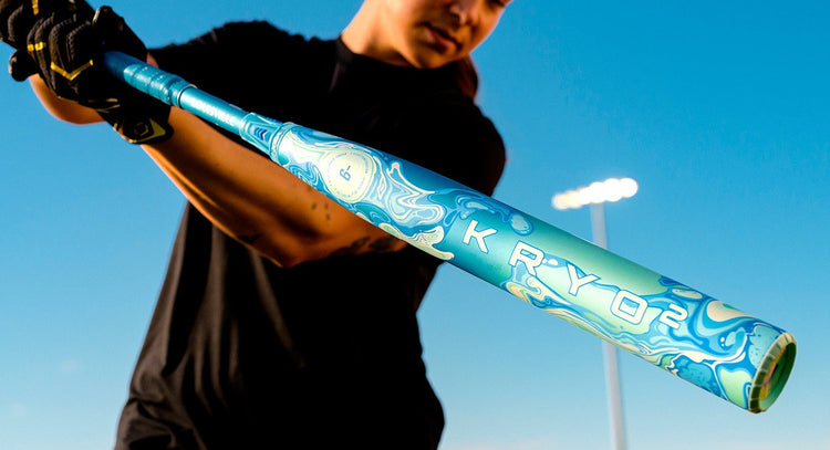 A person in black swings a colorful blue and white baseball bat labeled KRYOZ outdoors under a clear blue sky, with bright lights visible in the background.