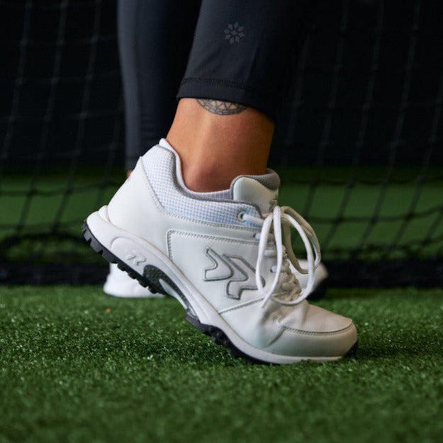 A person wearing white athletic shoes and black leggings stands on artificial turf, with one foot raised in front of a black net.