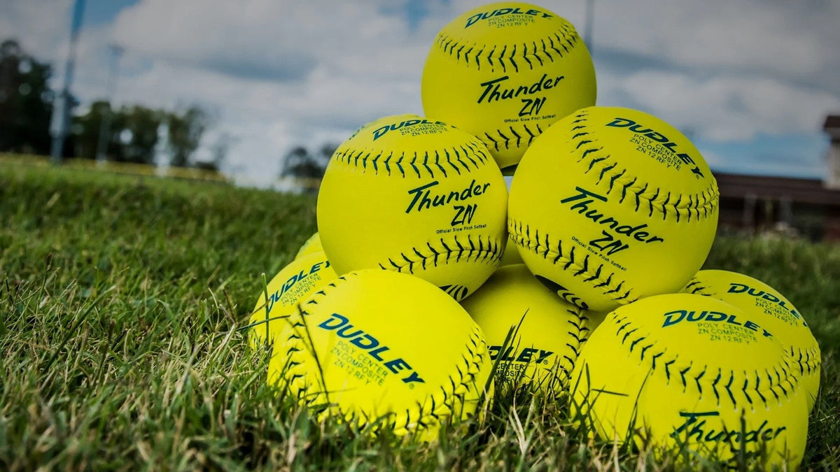 A pyramid stack of bright yellow softballs with “Dudley Thunder ZN” branding sits on green grass under a partly cloudy sky, with a blurred background of trees and a building.