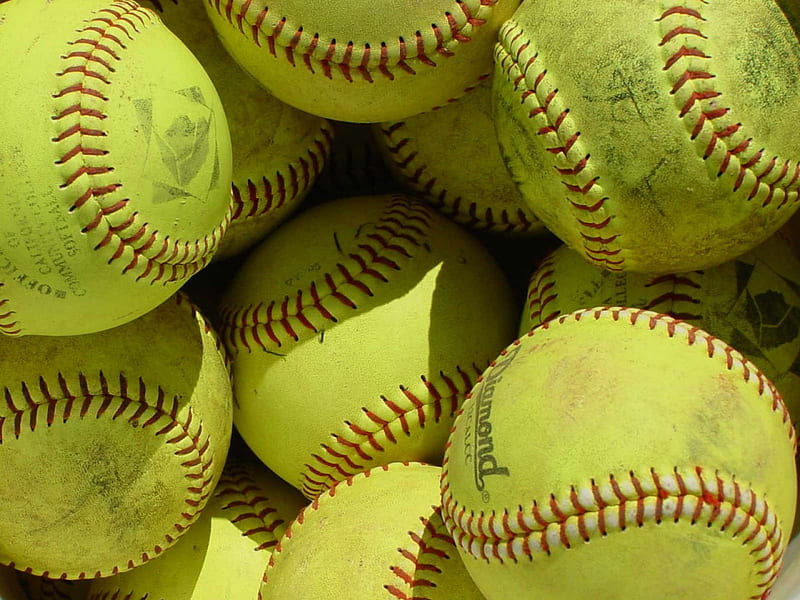 A close-up view of a pile of yellow softballs with red stitching, some showing signs of wear and visible printed logos or text.
