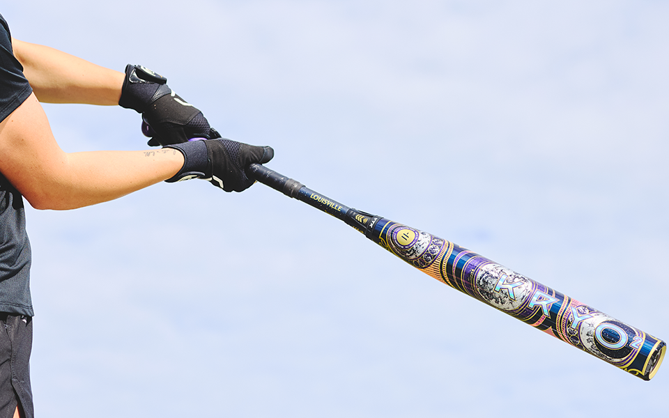 Wearing black gloves and shirt, a person grips the 2027 Louisville Slugger Kryo2 Empress Fastpitch Softball Bat, ready to swing against a bright, blurred background.