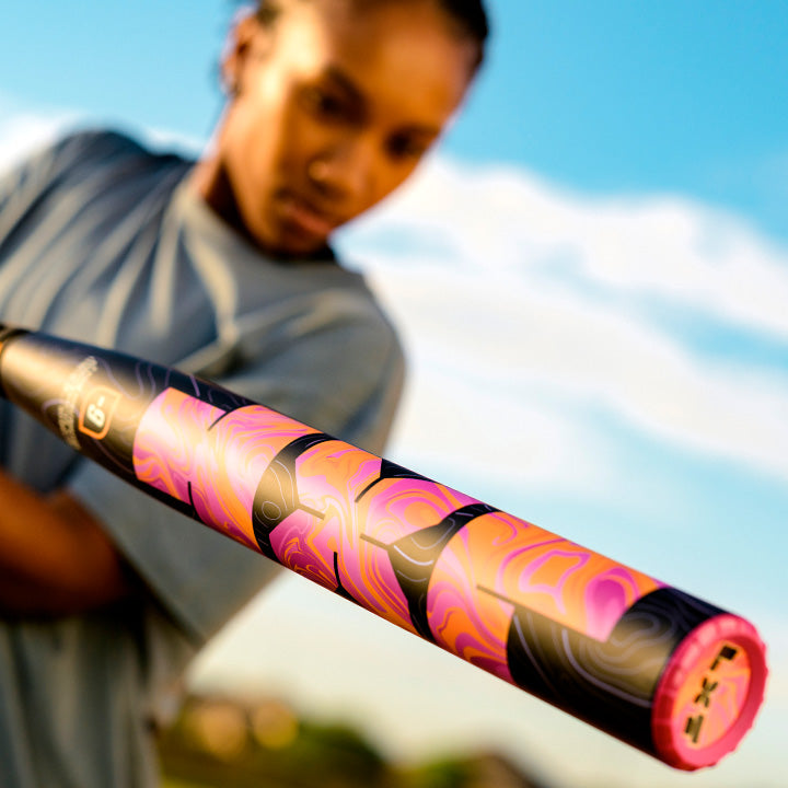 A person in a gray shirt holds a colorful, swirling pink and orange Louisville Slugger 2027 LXT (-10) Fastpitch Softball Bat (WBL4277010) with a composite barrel toward the camera, set against a blue sky background.