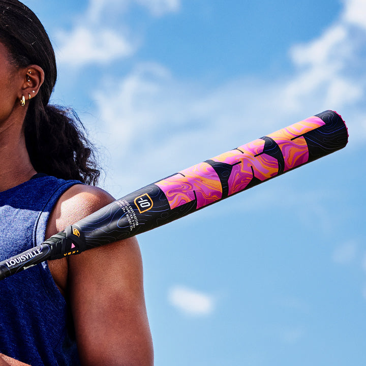 A person holding a vibrant Louisville Slugger LXT (-10) Fastpitch Softball Bat (2027, WBL4277010) with a composite barrel over their shoulder stands outdoors beneath a blue sky dotted with clouds. Only part of their upper body and face are visible, drawing attention to the bat’s balanced swing weight.