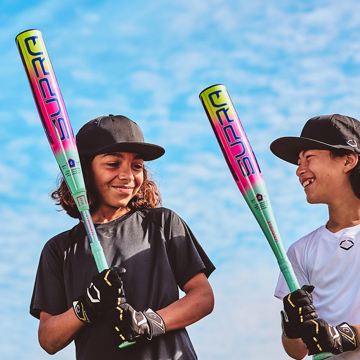 Two kids in black athletic gear and caps smile at each other, holding vibrant 2027 Louisville Slugger Supra (-10) 2 3/4" USSSA Baseball Bats (WBL4251010), standing outdoors beneath a bright blue sky with scattered clouds.