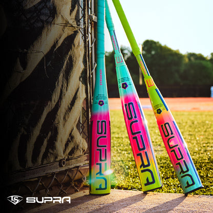 Three vibrant Louisville Slugger 2027 Supra JBB (-10) USSSA bats with gradient designs lean against a black net on a sunny field, lush grass and trees behind them. The Louisville Slugger logo appears in the lower left corner.