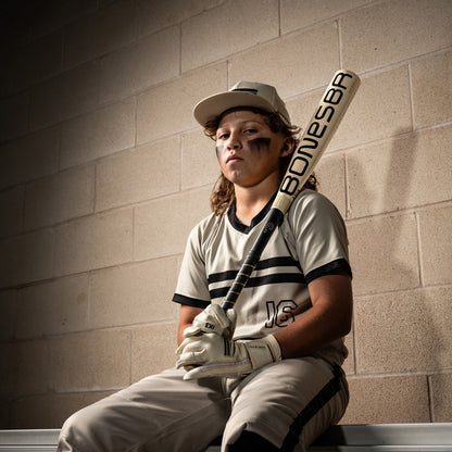 A young baseball player with long hair, eye black, gloves, and a cap sits on a bench against a brick wall, holding a Warstic 2026 Bonesaber Hybrid (-10) USA Baseball Bat (MBBSH25UBWH10) upright.