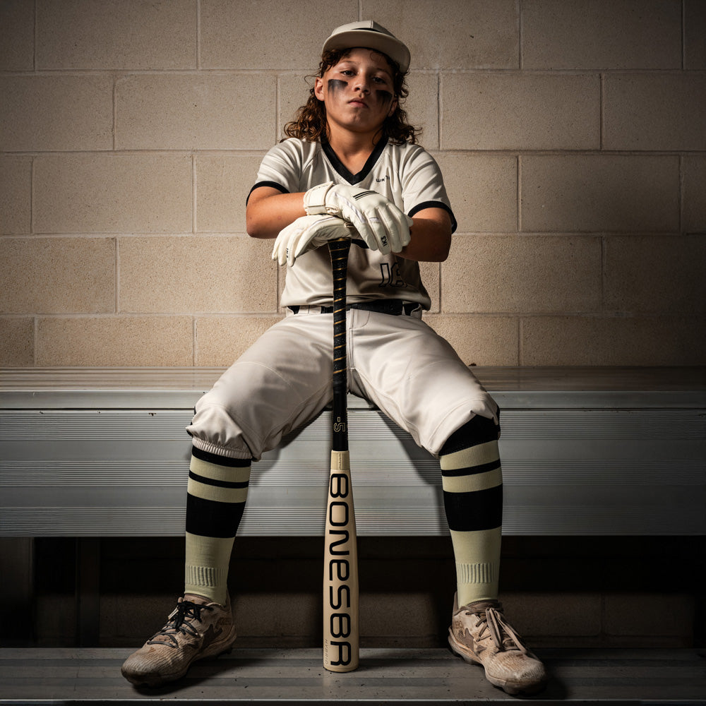 A girl sits on a bench holding a Warstic 2026 Bonesaber Hybrid (-10) USA Baseball Bat (MBBSH25UBWH10), her grip confident as she gazes out at the field.