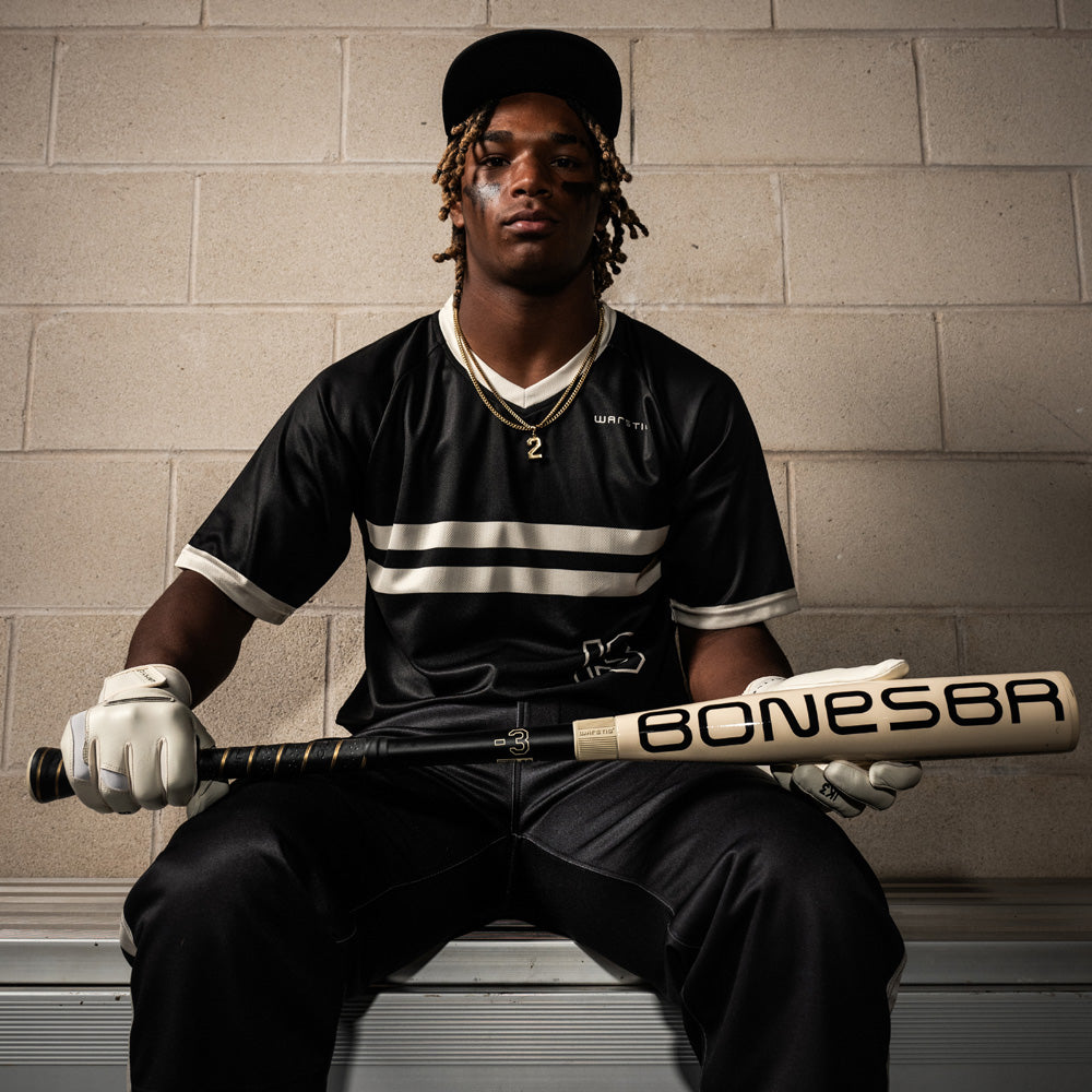A young man in a black sports uniform and white gloves sits on a bench holding a Warstic 2025 Bonesaber Hybrid (-3) BBCOR Baseball Bat (MBBSRHB25WH3) across his lap, with a neutral expression, against a tan brick wall.