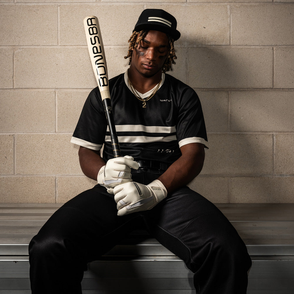A baseball player in a black and white uniform sits on a bench, holding a Warstic 2025 Bonesaber Hybrid (-3) BBCOR Baseball Bat (MBBSRHB25WH3). He wears a cap, gloves, and necklace, looking down with focus. A light brick wall is behind him.
