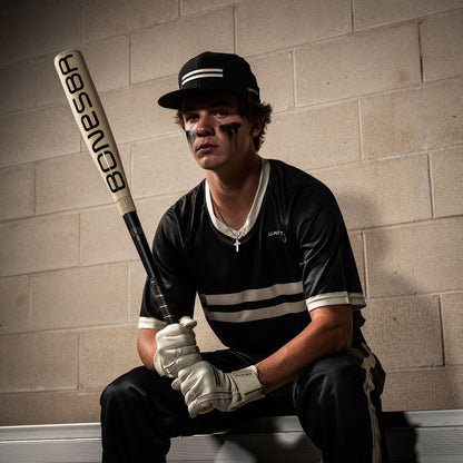 A baseball player in a black uniform and cap sits on a bench holding the 2025 Warstic Bonesaber Hybrid (-3) BBCOR Baseball Bat (MBBSRHB25WH3). He wears white gloves, a cross necklace, and black eye paint against a beige brick wall.