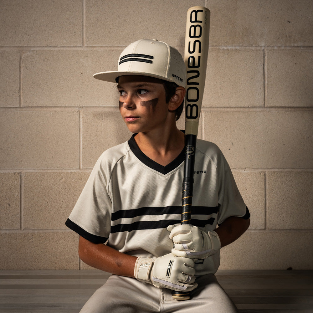 A young boy in a white baseball uniform sits on a bench, holding the 2026 Warstic Bonesaber Hybrid (-10) 2 5/8" USA Baseball Bat (MBBSH25UBWH10) vertically, looking to the side against a light brick wall background.