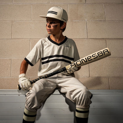 A young player in a white uniform with black stripes sits on a bench holding the 2026 Warstic Bonesaber Hybrid (-10) 2 5/8" USA Baseball Bat (MBBSH25UBWH10). He wears a cap, batting gloves, and black eye paint. A beige brick wall is behind him.