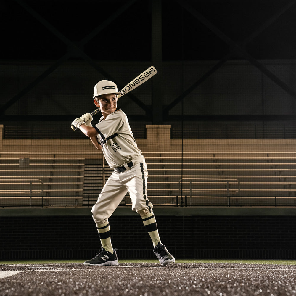 A young baseball player in a white uniform and helmet stands ready to bat on the field, gripping a Warstic 2026 Bonesaber Hybrid (-10) 2 5/8" USA Baseball Bat (MBBSH25UBWH10). Empty bleachers appear in the background.