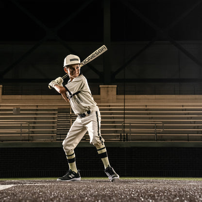 A young baseball player in a white uniform and helmet stands ready to bat on the field, gripping a Warstic 2026 Bonesaber Hybrid (-10) 2 5/8" USA Baseball Bat (MBBSH25UBWH10). Empty bleachers appear in the background.