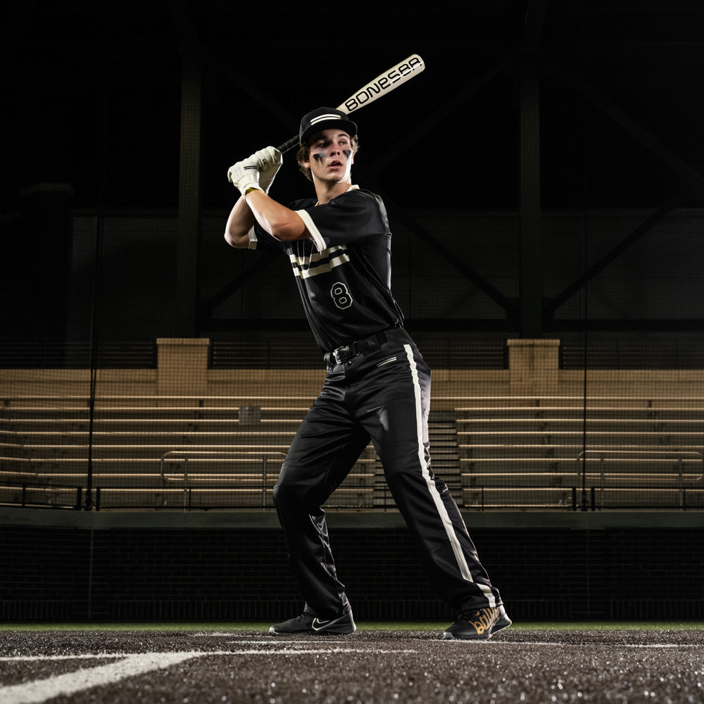 A baseball player in a black uniform stands ready to bat, gripping a Warstic 2025 Bonesaber Hybrid (-3) BBCOR Baseball Bat (MBBSRHB25WH3), with empty bleachers visible in the background.