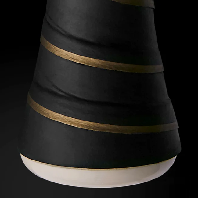 A close-up of a black vase with a matte finish, featuring diagonal gold stripes wrapped around its surface. The base of the vase is a contrasting smooth white.