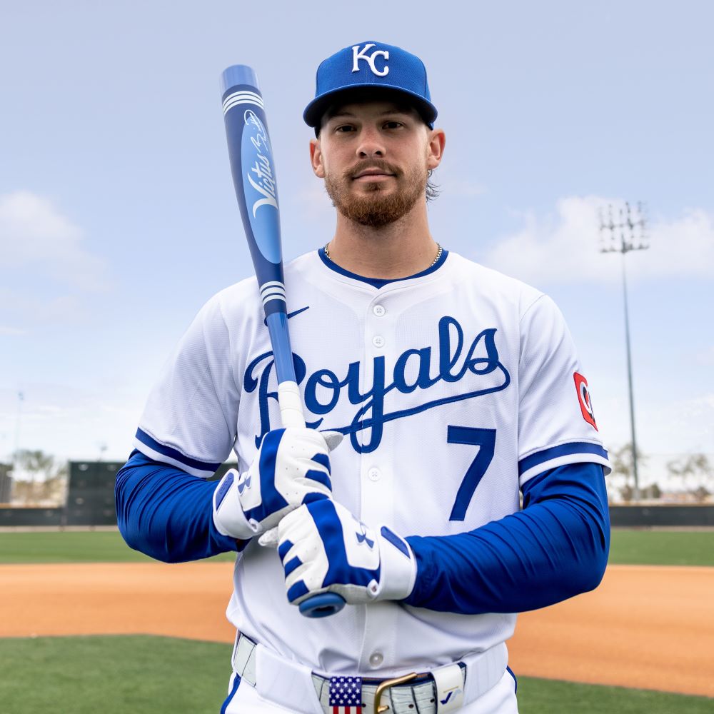 A Kansas City Royals player with number 7 stands on the field in a blue cap and gloves, holding the Victus 2025 Vibe Pro-Crayon Bobby Witt Jr (-5) 2 3/4" USSSA Baseball Bat (VSBVIB5C-BW), under clear skies and stadium lights.