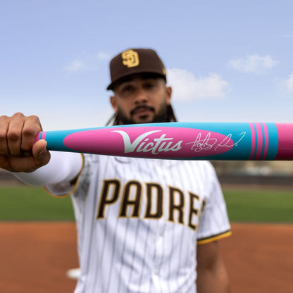 A Padres player holds a teal and pink 2025 Victus Vibe Pro-Crayon Fernando Tatis Jr (-8) USSSA bat with a signature toward the camera. The one-piece aluminum bat’s vibration reducing knob is sharply focused, the player's face blurred.