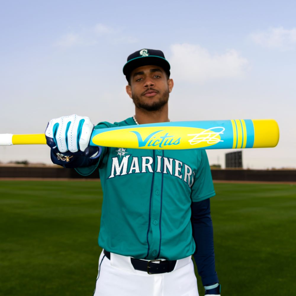 A baseball player in a Mariners uniform poses on the field, holding the Victus 2025 Vibe Pro-Crayon Julio Rodriguez (-5) USSSA Baseball Bat (VSBVIB5C-JR), featuring a bright yellow and blue design, knob for reduced vibration, and wearing cap and gloves.