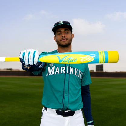 A baseball player in a Mariners uniform poses on the field, holding the Victus 2025 Vibe Pro-Crayon Julio Rodriguez (-5) USSSA Baseball Bat (VSBVIB5C-JR), featuring a bright yellow and blue design, knob for reduced vibration, and wearing cap and gloves.