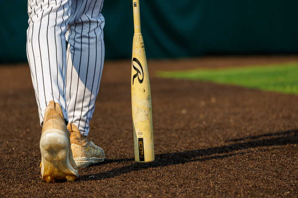 A baseball player in tan cleats and white pinstripe pants stands on a dirt field beside a yellow 2025 Rawlings Icon (-3) BBCOR Baseball Bat: RB5I3 (DEMO), with green grass visible in the background.