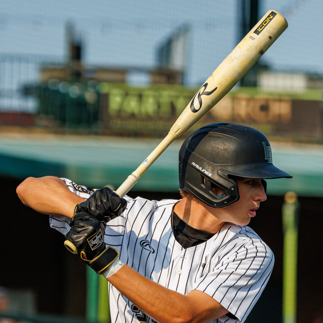 A baseball player in a black helmet and pinstriped uniform holds a bat over his shoulder, preparing to swing. The background shows a stadium with blurred seats and fencing.
