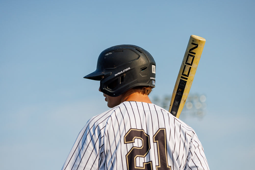 A baseball player in a black helmet and white pinstripe jersey with number 21 stands with a 2025 Rawlings Icon (-3) BBCOR Baseball Bat: RB5I3 (DEMO) by Rawlings resting on his shoulder against a clear blue sky.