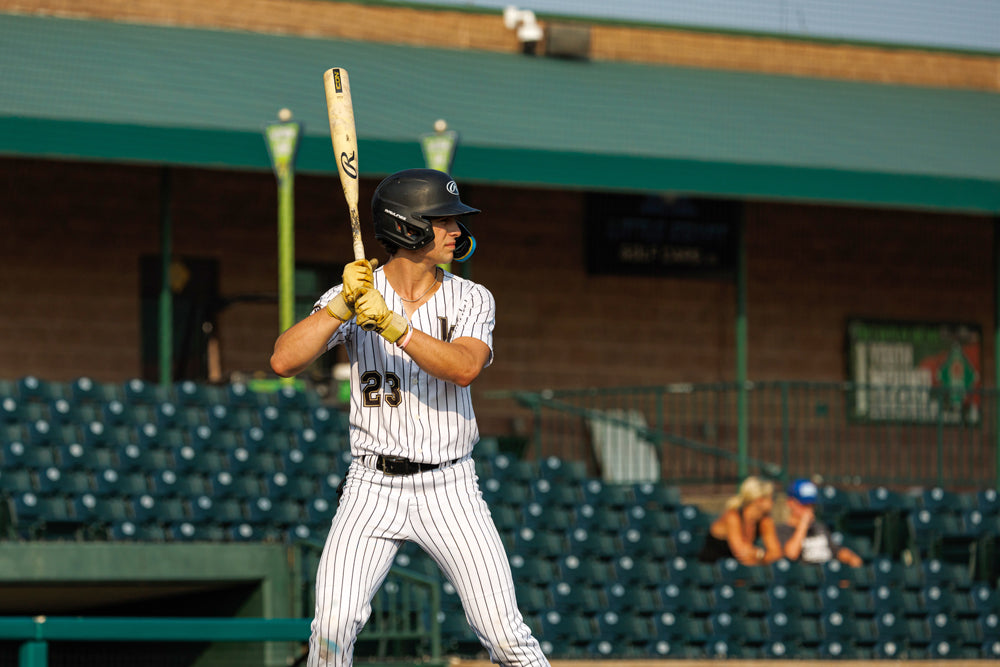 A baseball player in a black helmet and striped uniform stands ready to bat with the 2025 Rawlings Icon (-3) BBCOR Baseball Bat: RB5I3 (DEMO). Empty stadium seats and a few spectators can be seen in the background.