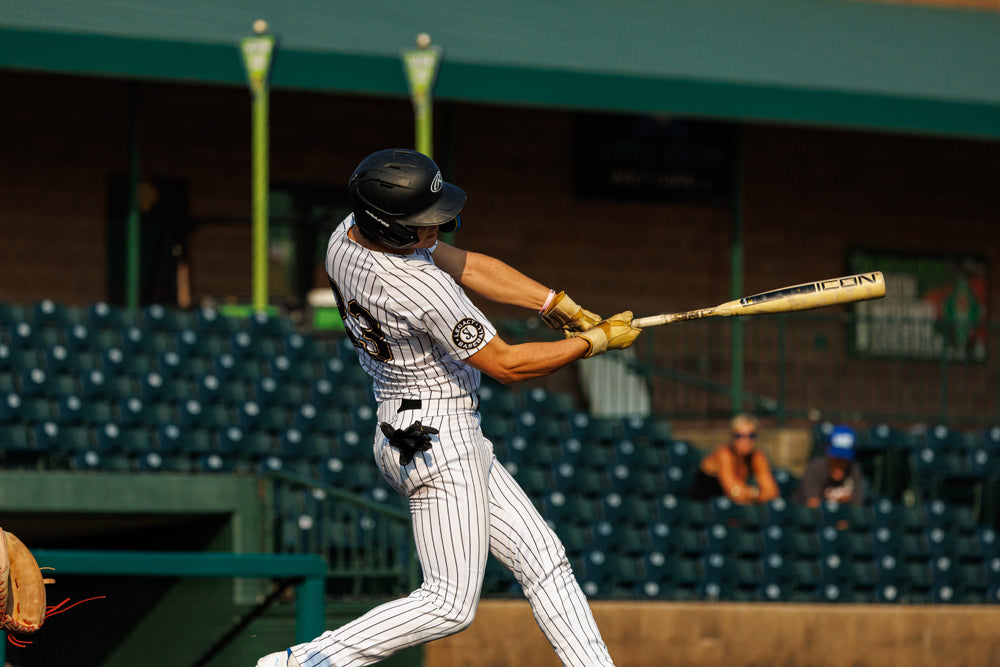 A baseball player in a white and black pinstripe uniform swings a 2025 Rawlings Icon (-3) BBCOR Baseball Bat: RB5I3 (DEMO) with helmet on, as empty stadium seats and a few spectators are visible in the background.