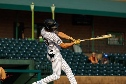 A baseball player in a white and black pinstripe uniform swings a 2025 Rawlings Icon (-3) BBCOR Baseball Bat: RB5I3 (DEMO) with helmet on, as empty stadium seats and a few spectators are visible in the background.