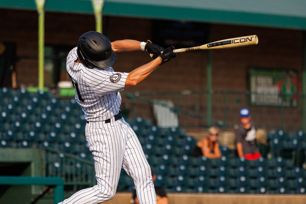 A baseball player in a black helmet and pinstripe uniform swings the 2025 Rawlings Icon (-3) BBCOR Baseball Bat: RB5I3 (DEMO) during a game, with empty stadium seats and a few spectators in the background.