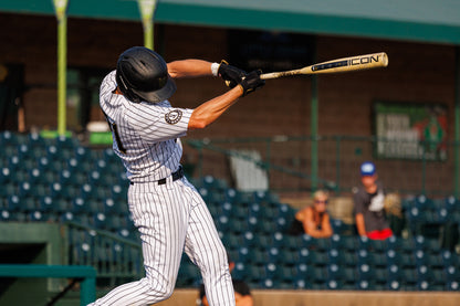 A baseball player in a black helmet and pinstripe uniform swings the 2025 Rawlings Icon (-3) BBCOR Baseball Bat: RB5I3 (DEMO) during a game, with empty stadium seats and a few spectators in the background.