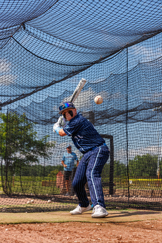 A young player in a blue pinstripe uniform readies a swing with a 2025 Rawlings Icon (-5) 2 3/4" USSSA Baseball Bat (USED) during batting practice, as the coach observes from behind the protective netting.