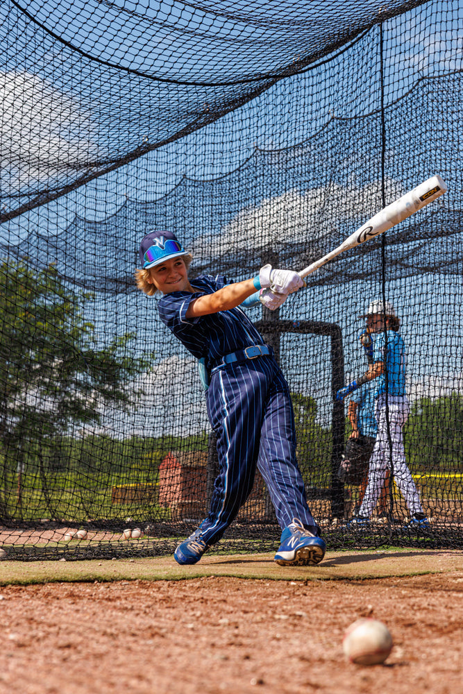 A young player in a blue pinstripe uniform swings a used Rawlings 2025 Icon (-8) 2 3/4" USSSA Baseball Bat (RUT5I8) in a batting cage, making contact with the ball while another player looks on under a partly cloudy sky.