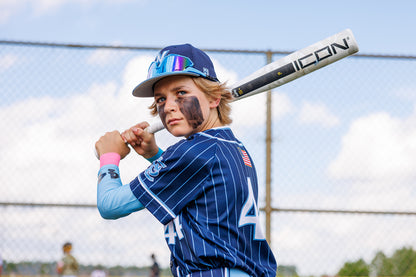 A young baseball player in a blue pinstripe uniform stands ready to bat, holding a used 2025 Rawlings Icon (-5) 2 3/4" USSSA Baseball Bat (RUT5I5) over his shoulder. He sports eye black, reflective sunglasses, and a chain-link fence background.