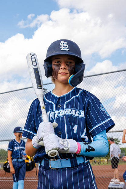 A young baseball player in a blue pinstripe uniform and helmet holds the 2025 Rawlings Icon (-5) 2 3/4" USSSA Baseball Bat (RUT5I5), with other players and a chain-link fence visible under a partly cloudy sky.