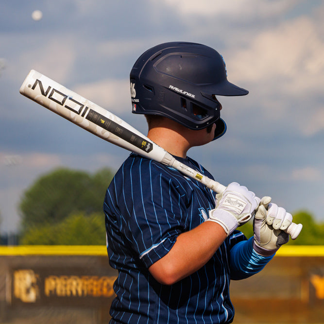 A baseball player in a navy pinstripe uniform and helmet stands ready to bat, holding a white bat over his shoulder on a sunny day, with a blurred field and sky in the background.