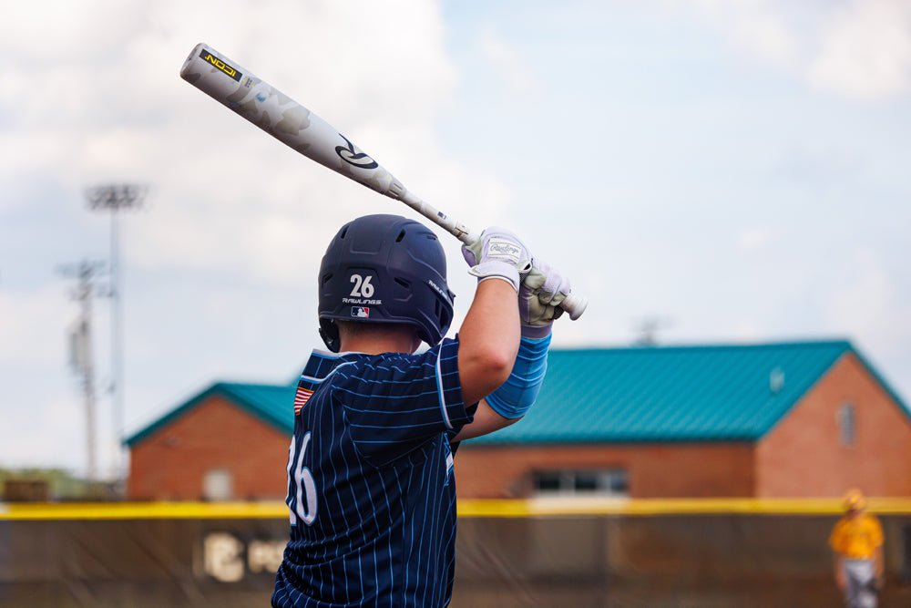 A baseball player in a navy pinstripe uniform and helmet readies to bat, holding the 2025 Rawlings Icon (-5) 2 3/4" USSSA Baseball Bat (RUT5I5). In the background, a brick building with a teal roof and another player can be seen.