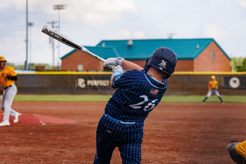 A baseball player in a blue pinstripe uniform swings a 2025 Rawlings Icon (-5) 2 3/4" USSSA Baseball Bat (RUT5I5) as players in yellow watch on a dirt field, with a brick building and fence visible in the background.